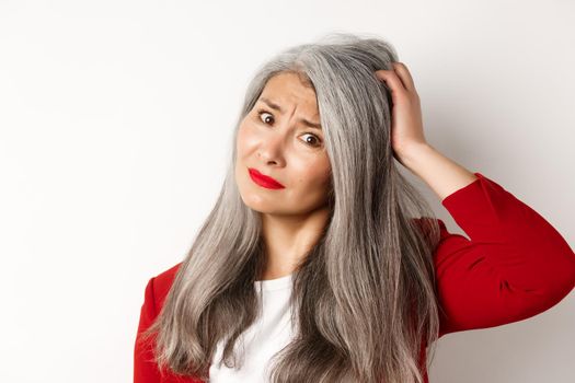 Close up portrait of confused asian female manager scratching head and looking questioned at camera, standing over white background.