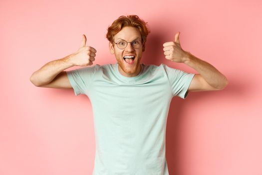 Amazed redhead guy, scream with joy and looking impressed and excited, showing thumbs up in approval, praise awesome promo offer, standing over pink background.