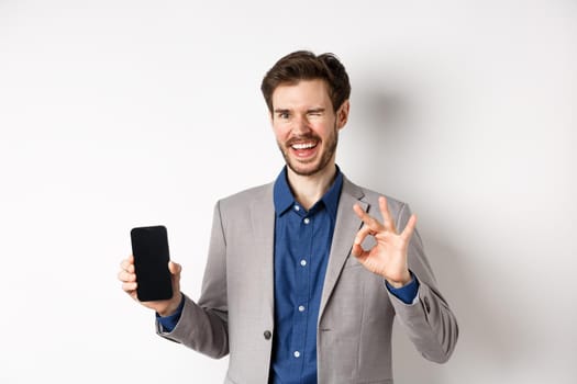 E-commerce and online shopping concept. Successful businessman making money on smartphone, showing empty phone screen and okay sign, winking happy at camera.