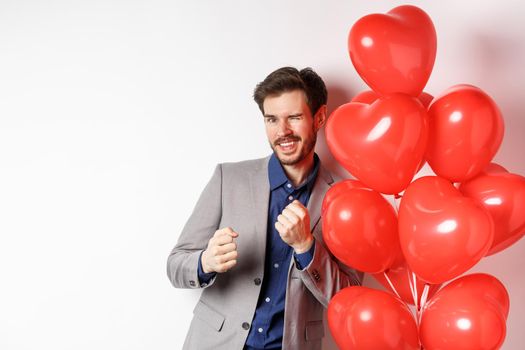 Handsome man dancing and smiling near Valentines day hearts balloon, winking at camera, getting dressed on romantic date on lovers holiday, white background.
