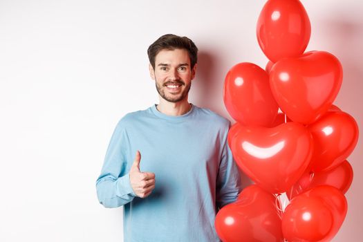 Valentines day and lovers concept. Happy young man showing thumbs up as standing with red hearts balloon, bring romantic gift on date, standing over white background.
