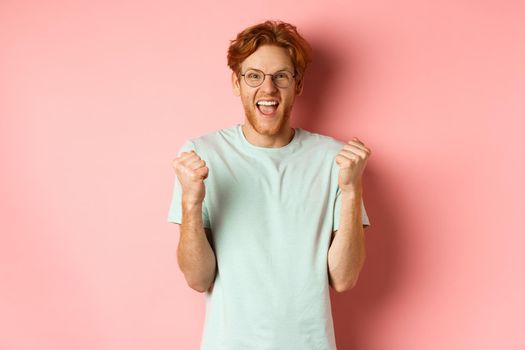 Excited young man winning prize, shouting with joy and triumph, making fist pump and saying yes, standing over pink background.