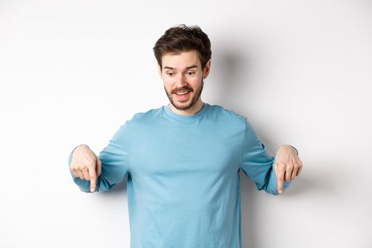 Excited and happy young man looking and pointing down at awesome deal, checking out promotion offer, standing on white background.