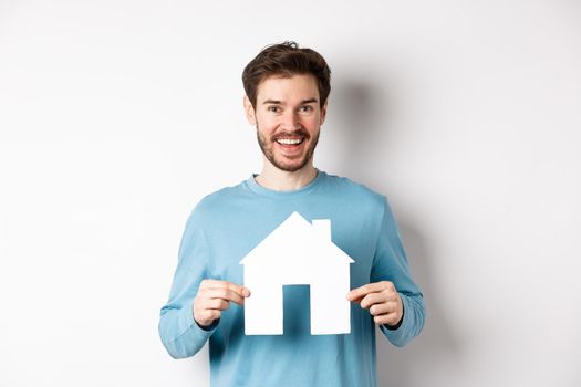 Real estate and insurance concept. Handsome modern man buying property, smiling and showing paper house cutout, standing over white background.