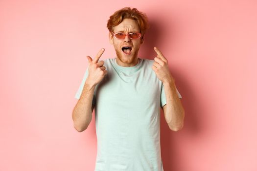Young handsome man with red hair and beard pointing fingers at sunglasses, showing new glasses, standing over pink background.