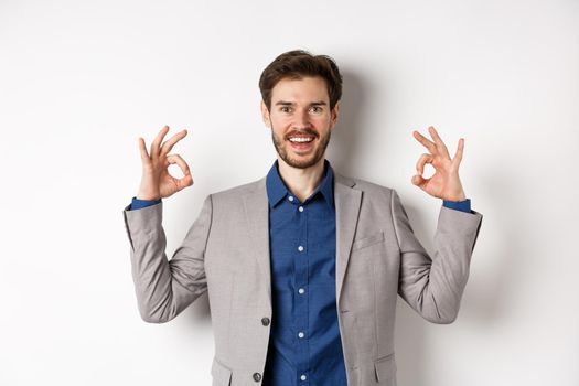 Good choice. Smiling businessman in suit showing okay signs and look happy, recommending good deal, like and approve your choice, standing satisfied on white background.