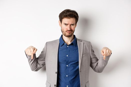 Disappointed bearded male entrepreneur frowning and pointing down with negative emotion, express dislike or bad opinion, standing on white background.