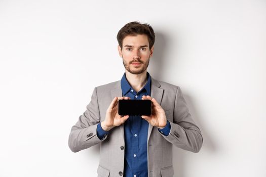 E-commerce and online shopping concept. Serious young man in suit showing empty smartphone screen horizontal, standing on white background.