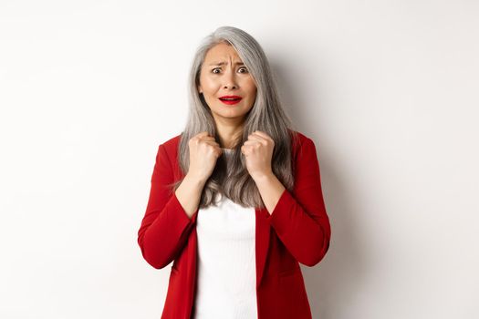 Business people. Scared mature asian woman looking terrified, trembling from fear, standing in red blazer over white background.