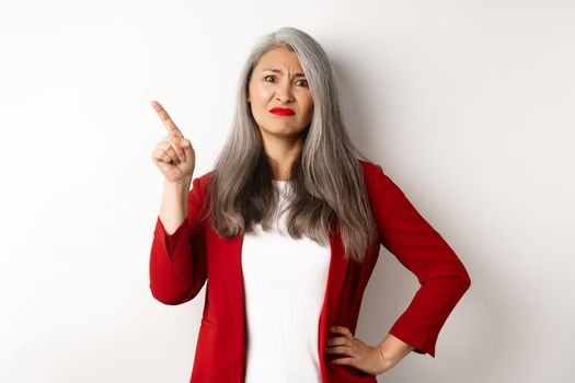 Skeptical asian businesswoman in red blazer grimacing, pointing upper left corner and looking dissatisfied, disapprove something, standing over white background.