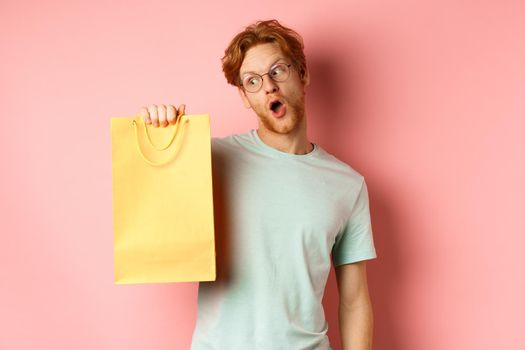 Funny handsome man with red hair, wearing glasses and t-shirt, holding and looking at yellow shopping bag, buying gifts during promo offer, standing over pink background.