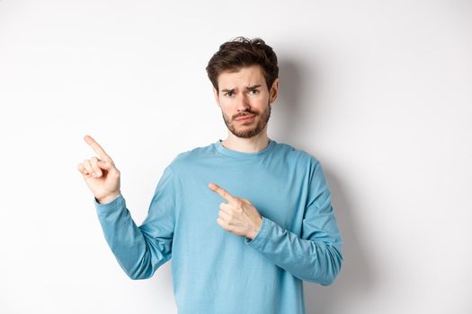 Skeptical young man with beard grimacing unsatisfied, frowning and pointing at upper left corner logo doubtful, standing upset over white background.