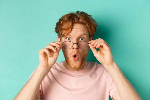 Surprised young man with red hair, checking out something cool, take-off glasses and saying wow impressed, standing over turquoise background.