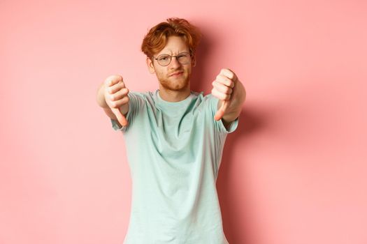 Disappointed young man in glasses, with messy red haircut, showing thumbs down and grimacing unsatisfied, express dislike, standing over pink background.