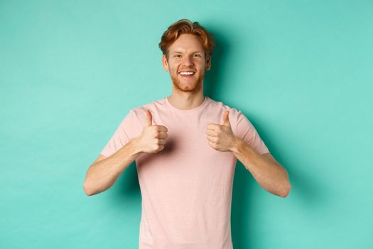 Cheerful bearded man with red hair showing thumbs-up, like and approve something, praising promo, standing against turquoise background.
