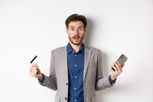 Online shopping. Confused man in suit spread hands sideways, holding plastic credit card with mobile phone and shrugging cueless, standing on white background.