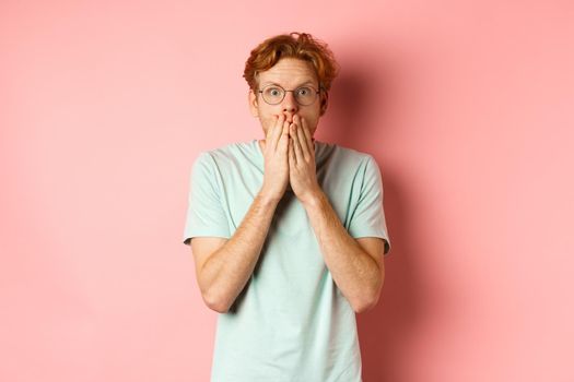 Shocked and speechless redhead guy covering mouth, staring at camera startled, standing in t-shirt against pink background.