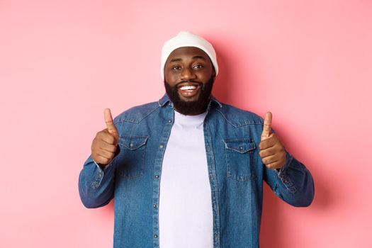 Happy african-american man showing thumbs-up in approval, like something good, praising great deal, standing over pink background.