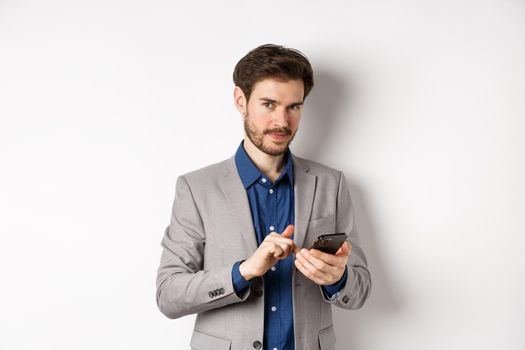 Handsome bearded male model in suit using mobile phone, smiling pleased at camera, white background.