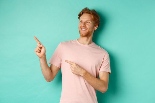 Handsome smiling man with red hair and beard looking delighted, pointing at upper left corner banner, standing in t-shirt over mint background.