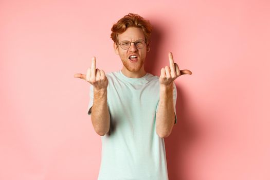 Arrogant and rude redhead man in glasses dont give a fuck, showing middle fingers at camera and frowning, standing over pink background.