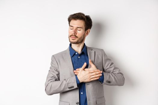 Romantic bearded man in stylish suit holding hands on heart, close eyes and remember something with tender feelings, standing nostalgic on white background.