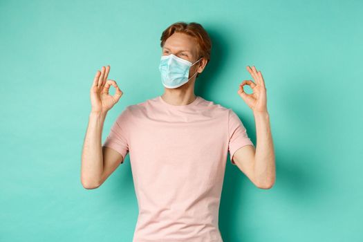 Covid-19, pandemic and lifestyle concept. Cheerful redhead man in medical mask showing okay signs in approval, like and praise product, looking left at copy space.