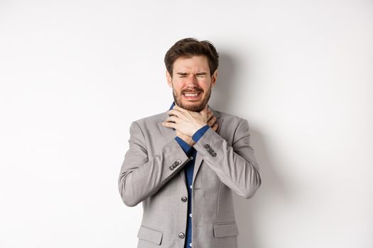 Crying man in suit holding hands on neck and look distressed, feel sick with sore throat, standing on white background.