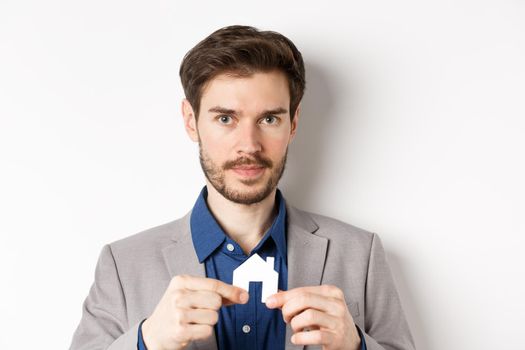 Real estate and insurance concept. Salesman realtor showing small paper house cutout and looking at camera, selling property, white background.