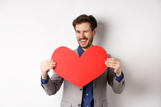 Charismatic young man winking and smiling, showing big red heart cutout for Valentines day date, say love you to lover, standing over white background.