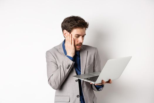 Shocked and worried businessman making mistake on laptop, looking at computer upset, standing on white background.