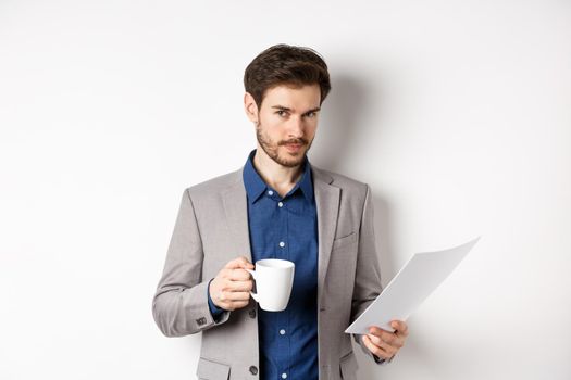 Businessman in suit reading documents papers and drinking coffee, smiling confident at camera, white background.