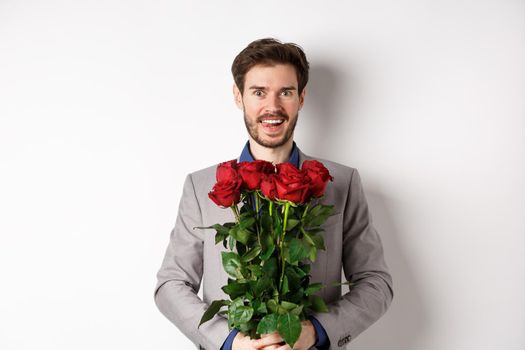 Excited handsome man in suit holding bouquet of roses for romantic date with lover, standing happy on Valentines day, white background.