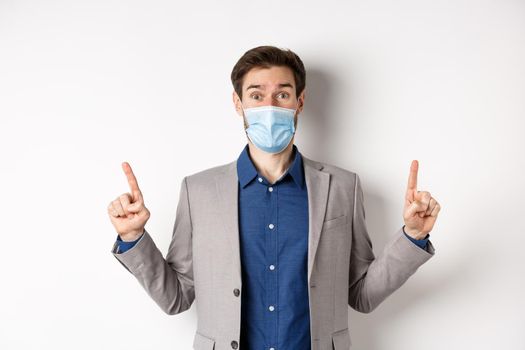 Covid-19, pandemic and business concept. Excited businessman in sterile medical mask and suit pointing fingers up, white background.