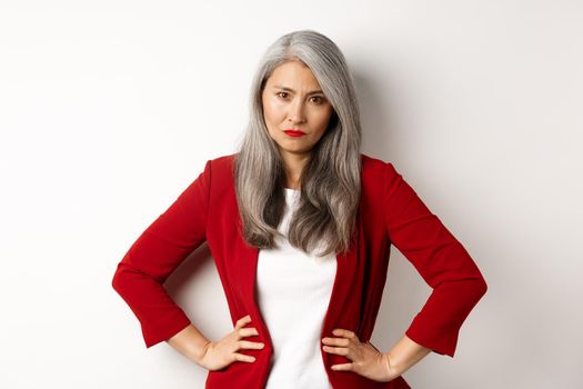 Angry asian senior businesswoman scolding employee, holding hands on waist and staring disappointed at camera, white background.
