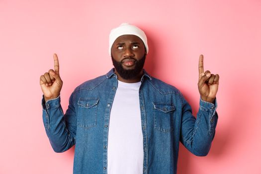 Concerned and hesitant Black man in beanie pointing fingers up, staring at top with skeptical face, standing over pink background.