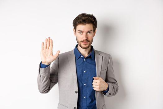 Businessman in formal suit raising hand up to say hello, contactless greeting, smiling friendly, standing on white background.