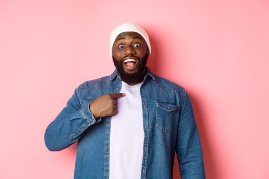 Surprised Black man in hipster beanie pointing at himself, staring at camera amazed, standing over pink background.