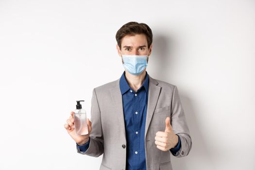 Covid-19, pandemic and business concept. Businessman in office suit and medical mask showing bottle of hand sanitizer and thumb up, recommend use antiseptic at work.