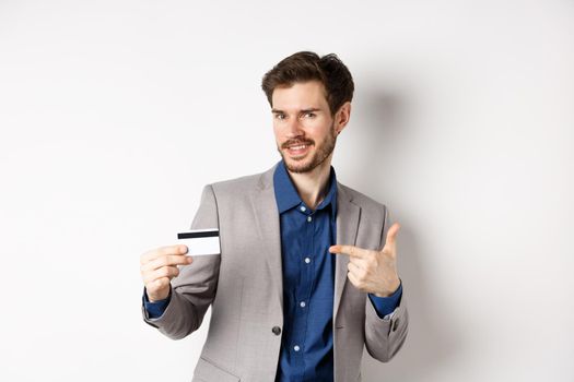 Successful male entrepreneur pointing at plastic credit card and smiling, recommending bank, standing on white background.