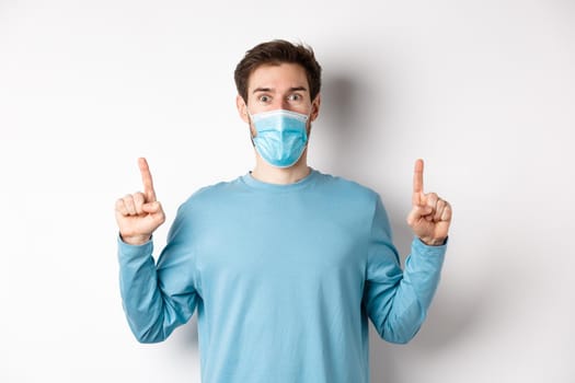 Coronavirus, health and quarantine concept. Excited smiling guy checking out special deal, pointing fingers up, stare amazed at camera, wear medical mask on white background.