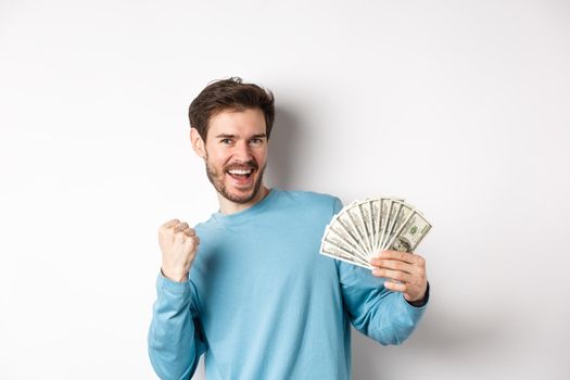 Happy man dancing with money, showing dollars and saying yes with satisfied smile, making fist pump gesture, standing over white background.