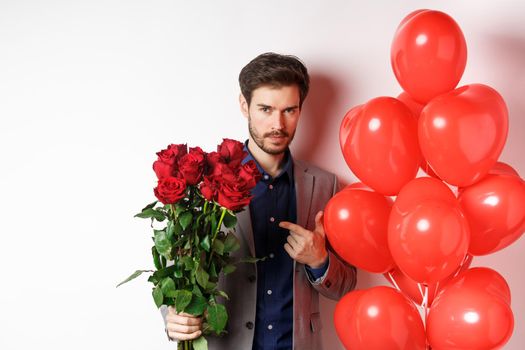Handsome man in suit going on romantic Valentines day date, pointing at bouquet of roses, standing near heart balloon over white background.