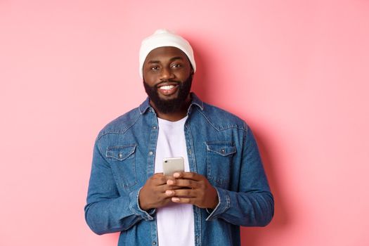 Handsome african-american guy using smartphone, smiling at camera, standing over pink background.