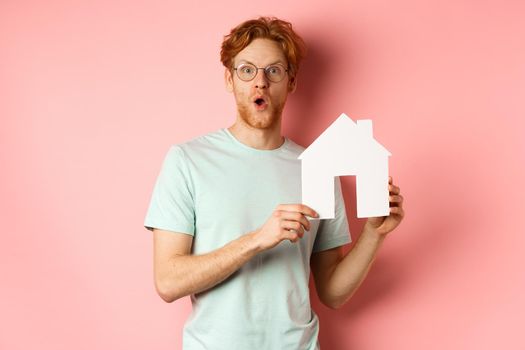 Real estate. Surprised young man with red hair and beard, wearing glasses and t-shirt, showing paper house cutout and looking impressed, saying wow, standing over pink background.