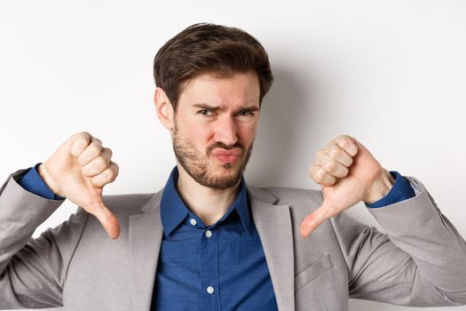 Close up of skeptical young man in suit showing thumbs down, dislike and judging bad product, negative emotion, white background.