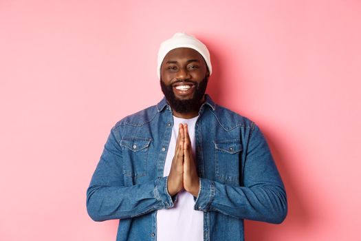 Happy smiling Black man saying thank you, holding hands in pray or namaste gesture, standing grateful against pink background.