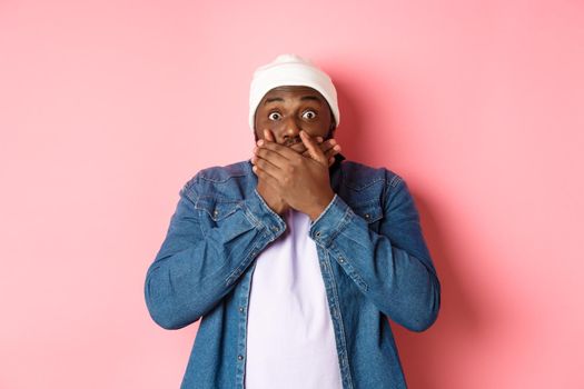 Shocked african-american man cover mouth and staring at camera, standing speechless against pink background.