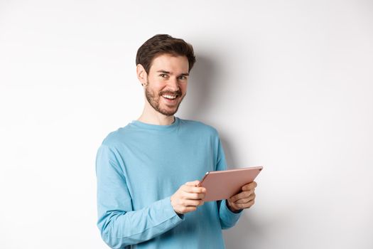 E-commerce and technology concept. Young caucasian man smiling at camera, holding digital tablet, standing over white background.