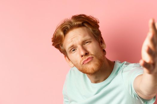 Funny young man with red messy hair taking selife with puckered lips and macho expression, view from smartphone camera, standing over pink background.